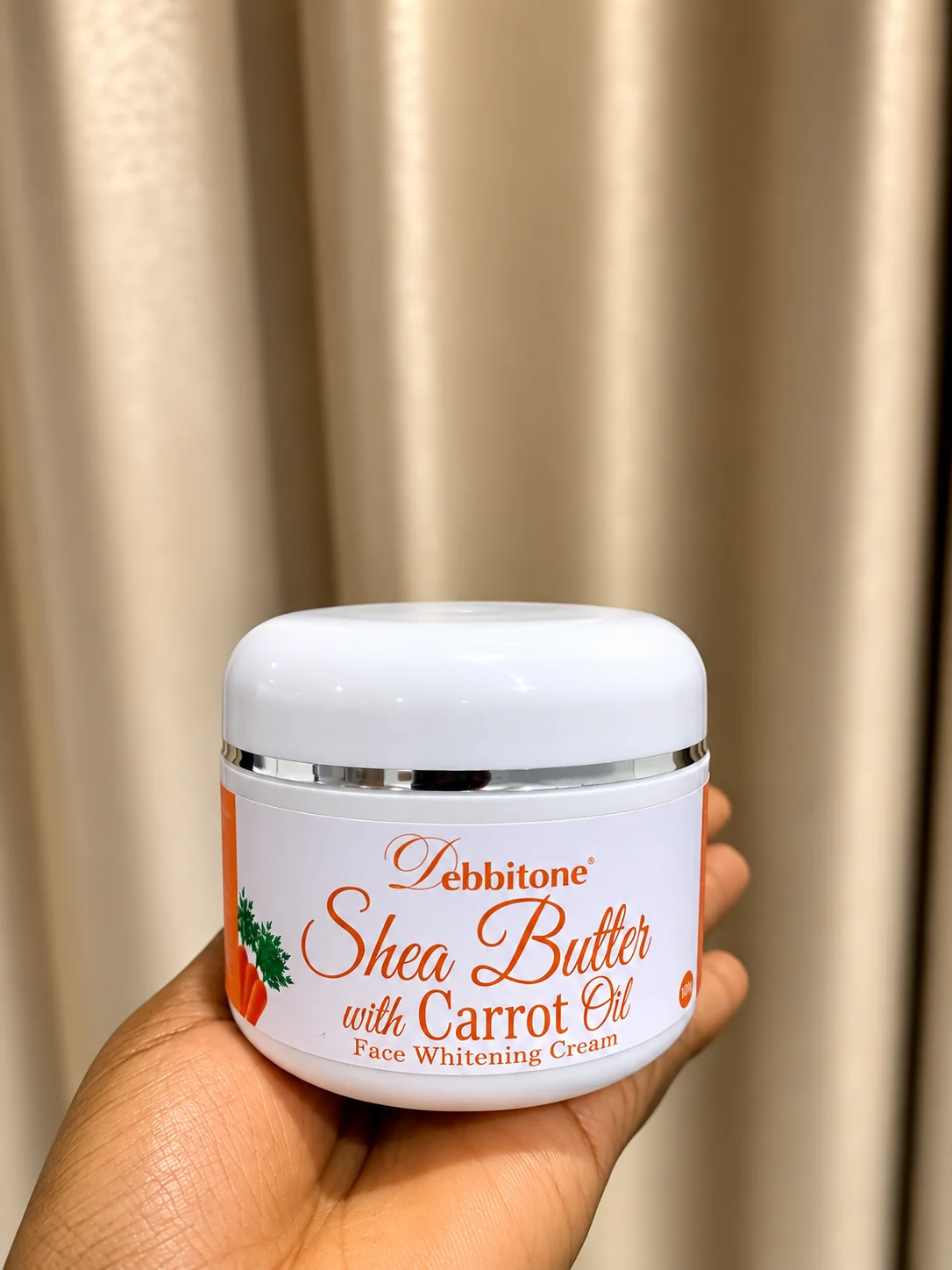 A 160g jar of Debbitone SheaButter Carrot Brightening Cream held in one hand
